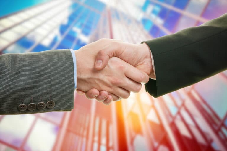 Symbolic picture of two men shaking hands in business suits front of a skyscraper.